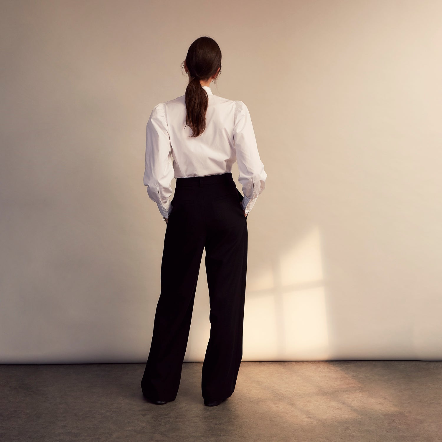 View 6 of product A woman stands in profile, looking down with hands in her pockets, wearing the Turnbull & Asser Turnbull and The Deck White Cotton Expressionist Shirt, paired with high-waisted black-and-white houndstooth pants.