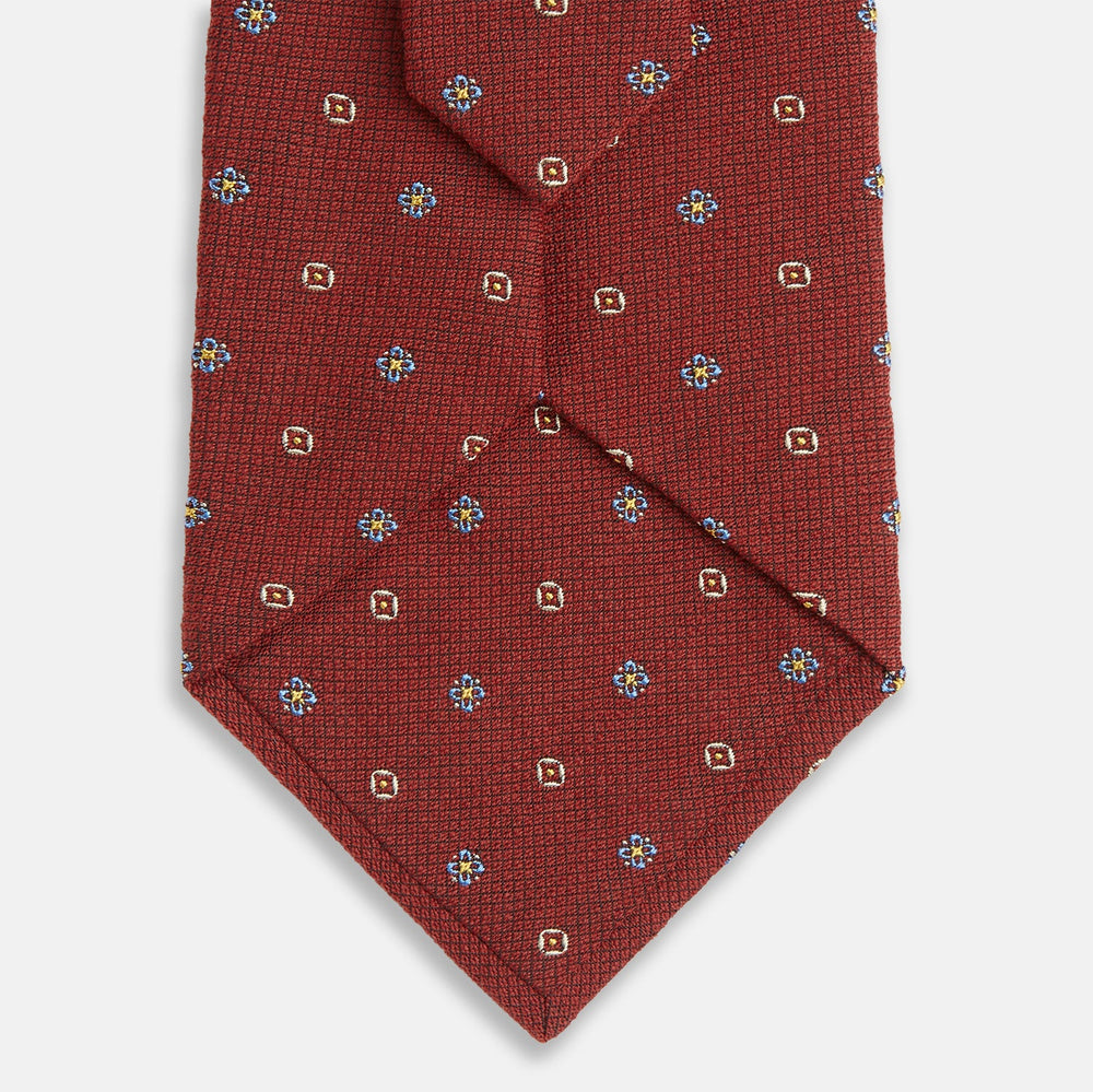 A Turnbull & Asser Red Motif Silk Tie featuring small blue and yellow floral designs with brown geometric shapes is displayed rolled up on a plain white background.