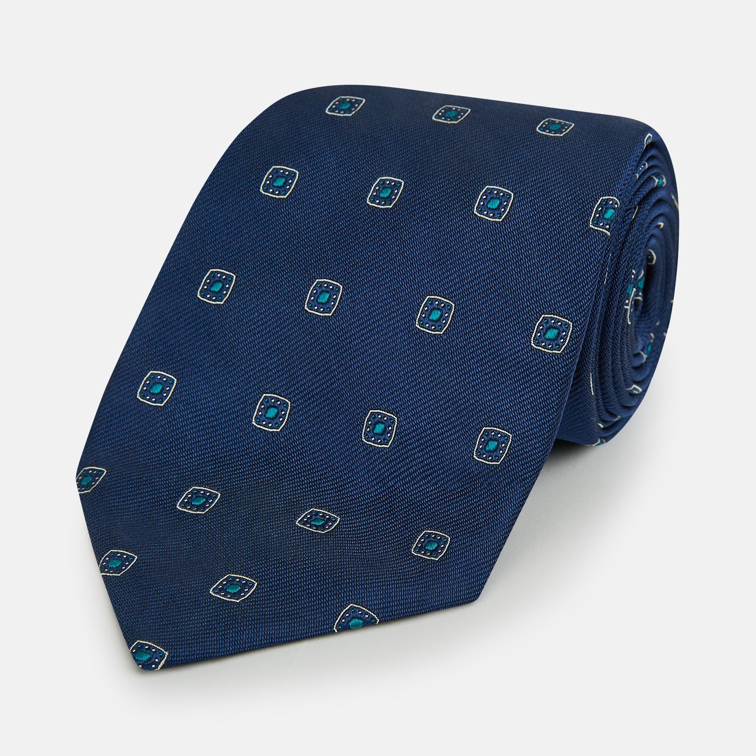 View 1 of product The Turnbull & Asser Navy Geometric Silk Tie showcases a repeating pattern of small light blue and white shapes on a navy base, neatly rolled and displayed against a white background.