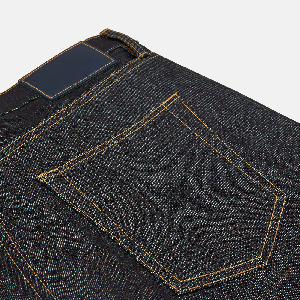 A pair of Turnbull & Asser Indigo Japanese Selvedge Denim Jeans in dark blue with rolled-up cuffs, yellow stitching, five pockets, and a button fly, laid flat on a white background.