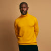 View 1 of product A man with short hair and a beard wears the Gold Aubrey Cashmere Roll Neck Jumper by Turnbull & Asser with dark pants, standing against a plain beige background and calmly looking at the camera, highlighting this stylish autumn sweater look.