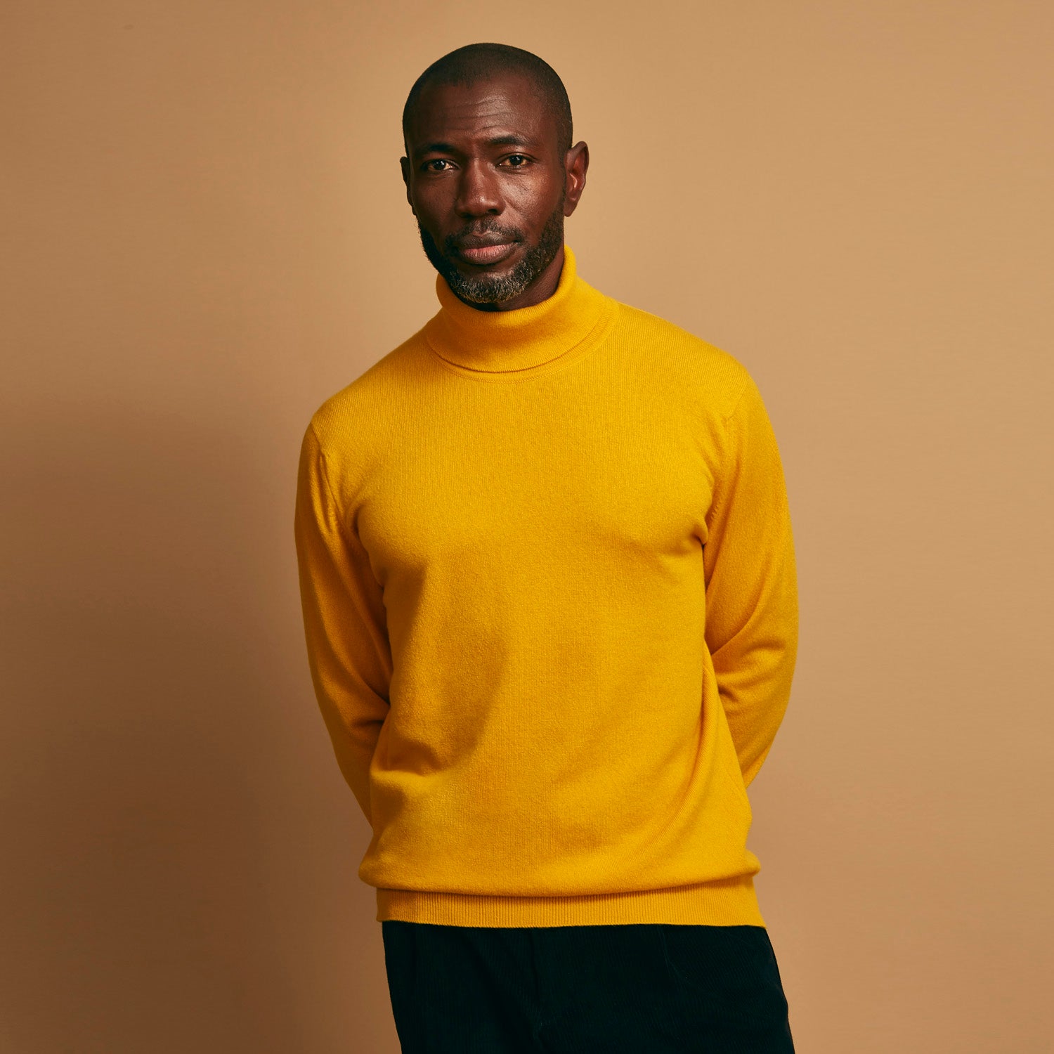 View 1 of product A man with short hair and a beard wears the Gold Aubrey Cashmere Roll Neck Jumper by Turnbull & Asser with dark pants, standing against a plain beige background and calmly looking at the camera, highlighting this stylish autumn sweater look.