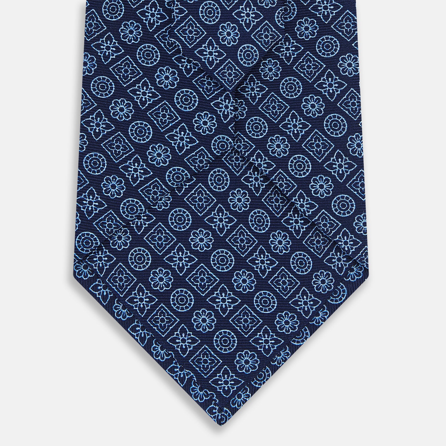 View 3 of product The Turnbull & Asser Dark Navy Floral Tile Pattern Printed Silk Tie, rolled up and displayed on a white background, features a blue geometric and floral pattern.