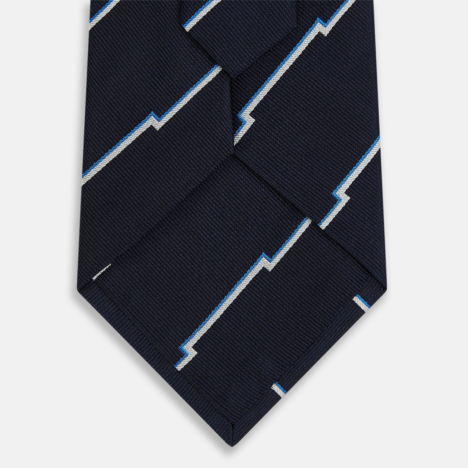 View 3 of product The Turnbull & Asser Navy Broken Shadow Stripe Silk Tie features British excellence with a light blue and white zigzag pattern and is shown rolled up on a plain white background.