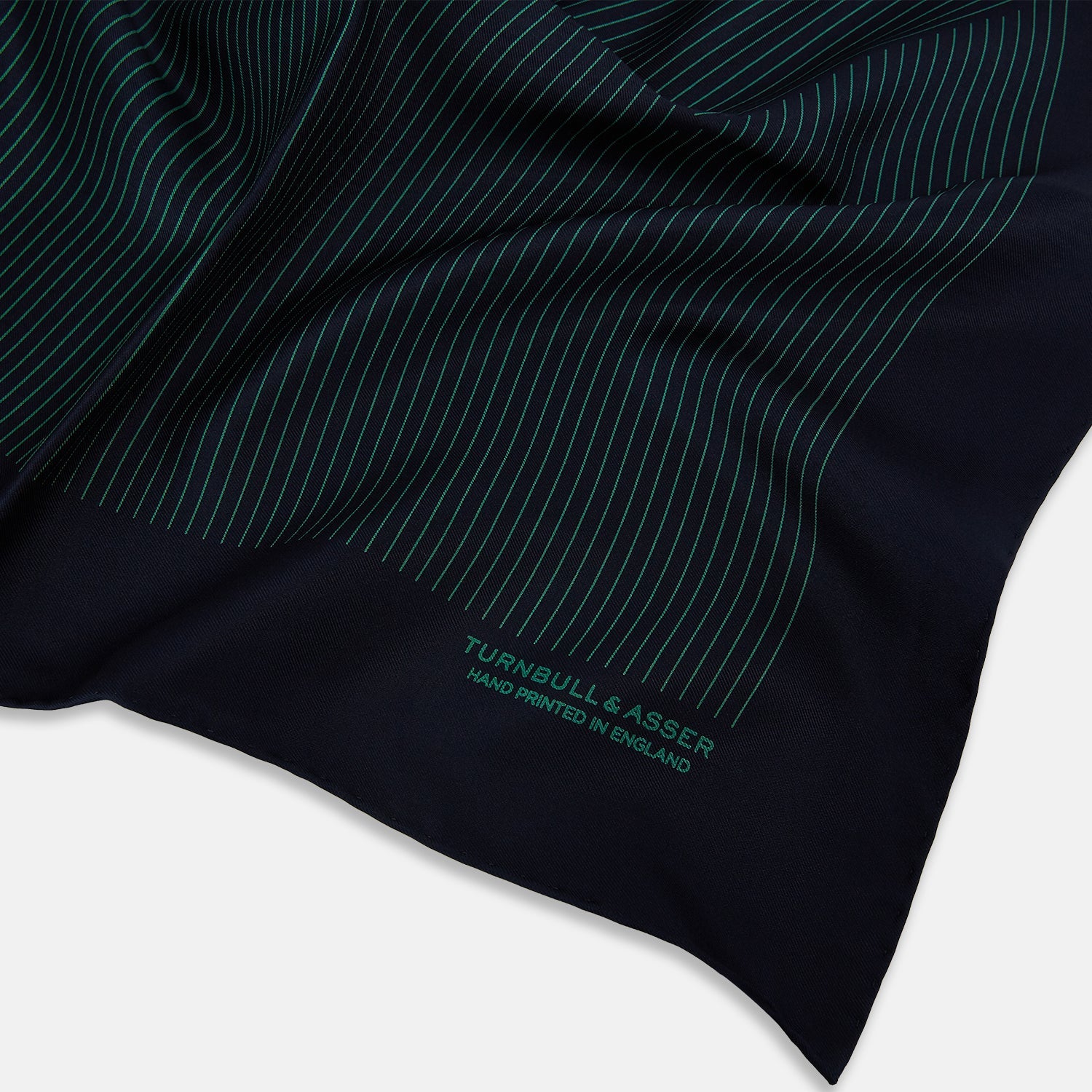 View 2 of product The Turnbull & Asser Navy and Hunter Green Stripe Silk Pocket Square features thin green stripes, hand-rolled edges, and "TURNBULL & ASSER HAND PRINTED IN ENGLAND" in green on the bottom right corner.
