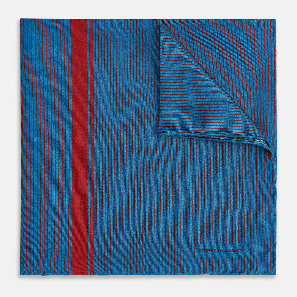 A blue silk pocket square by Turnbull & Asser, featuring thin black stripes, a striking vertical red stripe, and a turned-over corner that reveals the reverse side. The brand name is printed discreetly in one corner.