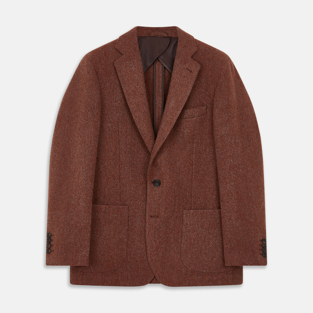 A man stands in a lush garden, hands in pockets and relaxed, wearing the Turnbull & Asser Red Lambswool Herringbone Bern Blazer, a striped dress shirt, patterned scarf, and dark trousers.
