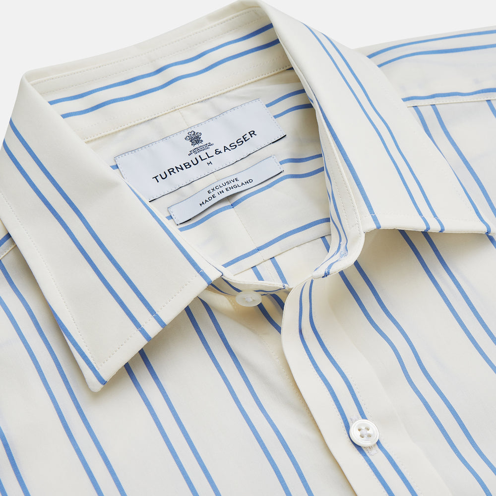 A man with short brown hair stands against a beige background in a Turnbull & Asser Blue and Off-White Stripe Cotton Weekend Fit Finch Shirt, featuring vertical stripes and a Derby collar, paired with dark pants. One hand is in his pocket; he has a neutral expression.
