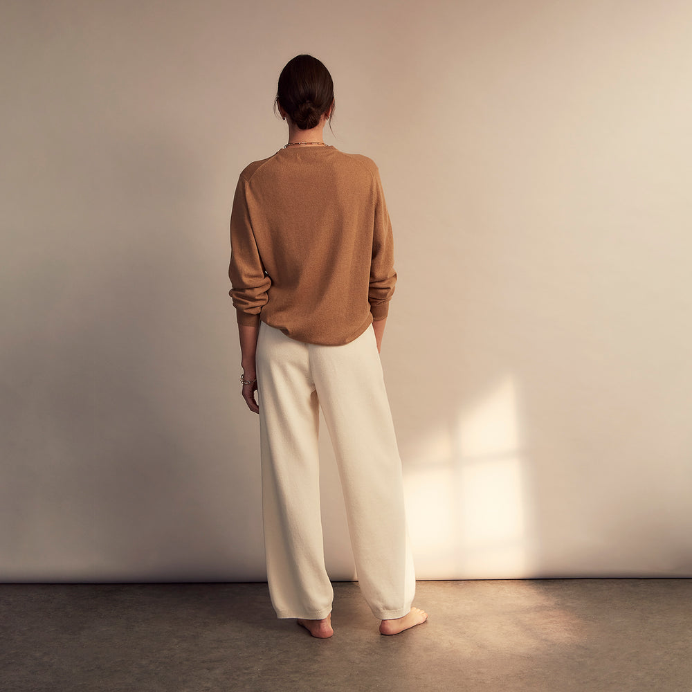 A woman stands barefoot indoors against a neutral wall, wearing Turnbull & Asser Women's Cream Cashmere Knitted Tabitha Bottoms and a light brown sweater. Soft sunlight casts a gentle shadow on the wall beside her.