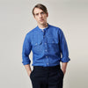 View 4 of product A man faces forward with a neutral expression, hands in his pockets, wearing the Turnbull & Asser Plain Indigo Linen Weekend Fit Shirt with a stand collar, styled with dark pants and black shoes against a plain light background.