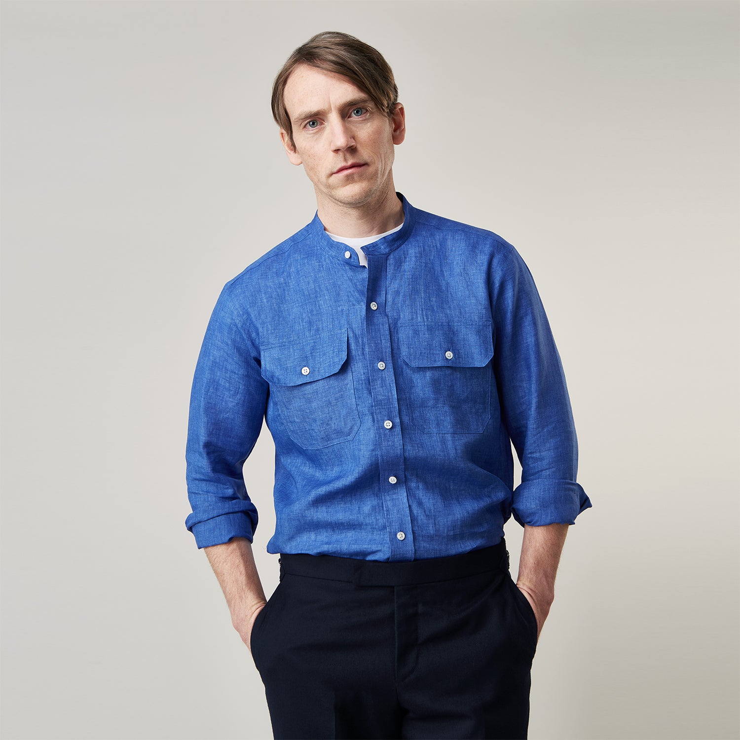 View 4 of product A man faces forward with a neutral expression, hands in his pockets, wearing the Turnbull & Asser Plain Indigo Linen Weekend Fit Shirt with a stand collar, styled with dark pants and black shoes against a plain light background.