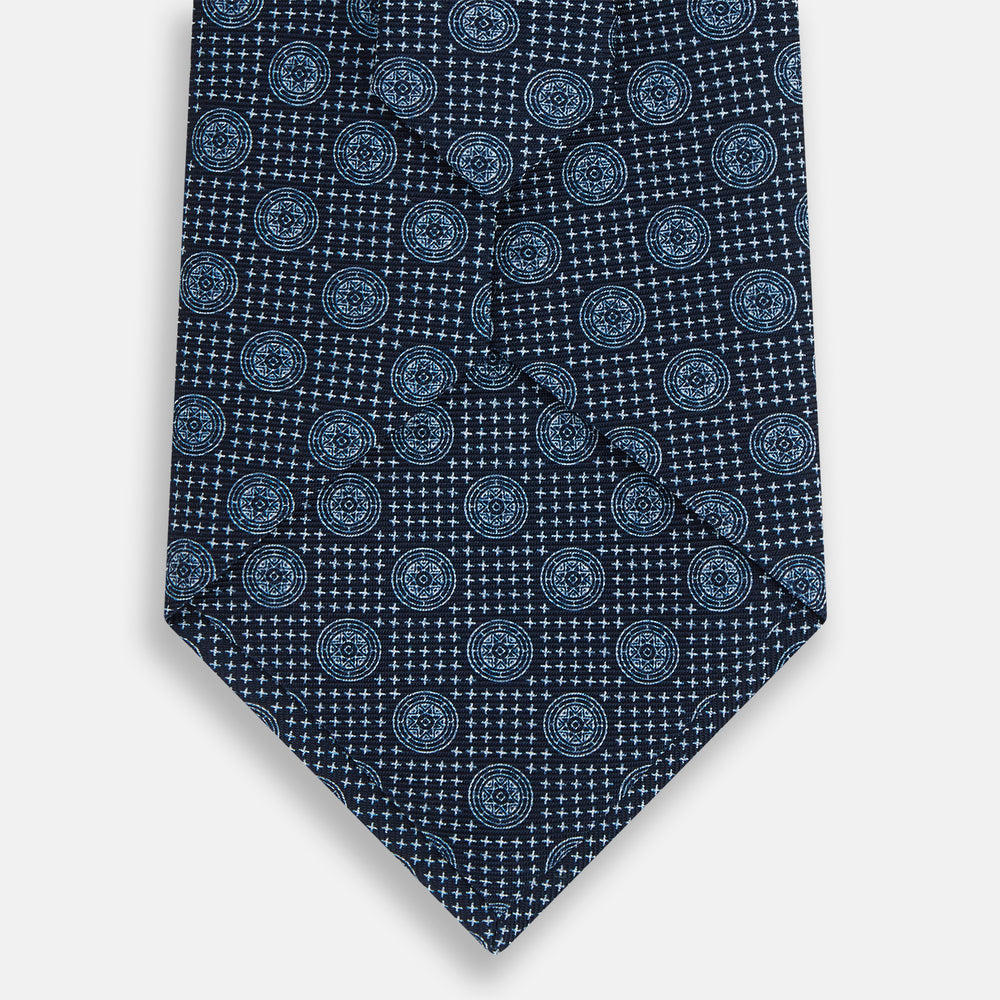 The Turnbull & Asser Navy Medallion Silk Tie is crafted from fine English silk, featuring light blue geometric medallions on a white background, and is rolled neatly with the pointed end facing forward.