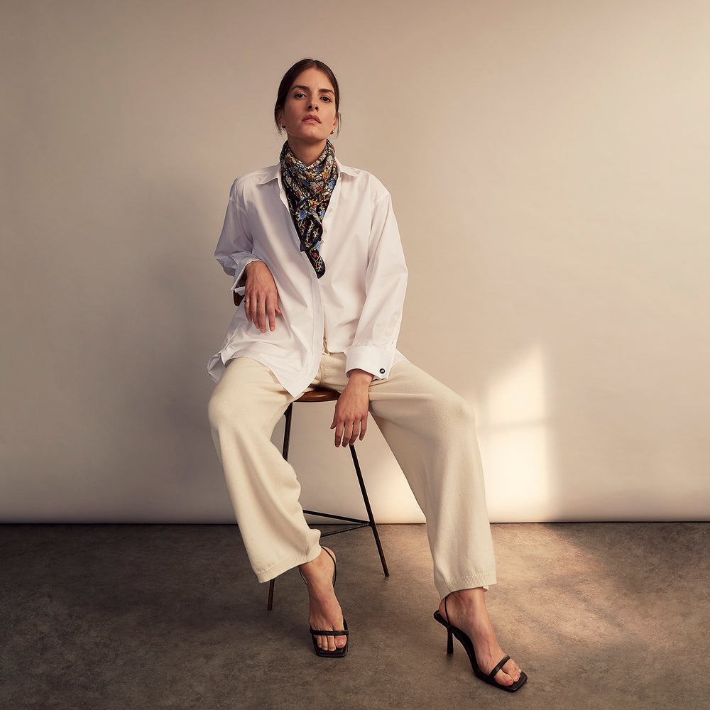 A woman with straight brown hair pulled back wears the Turnbull & Asser White Cotton Marlene Shirt, featuring classic menswear details, paired with high-waisted cream trousers against a neutral background.