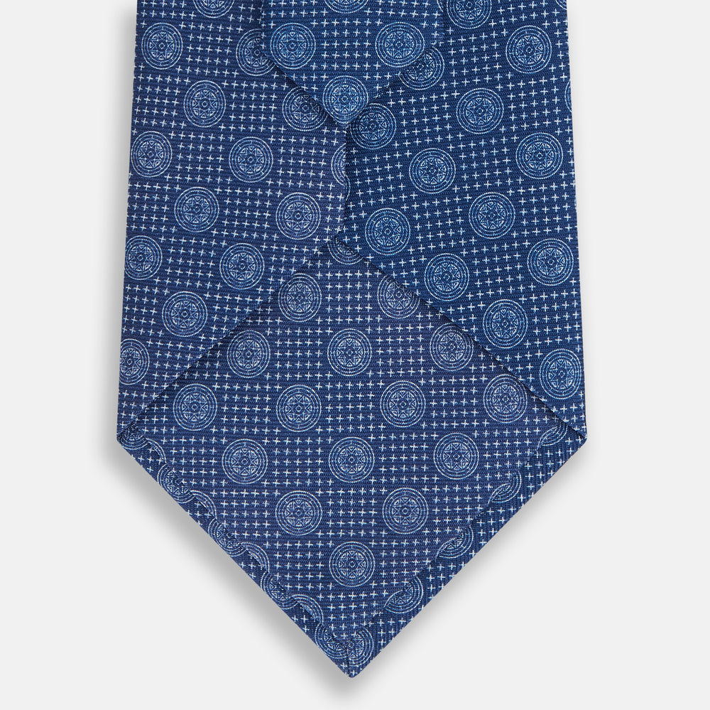 The Turnbull & Asser Azzurro Medallion Silk Tie, rolled up and displayed on a white background, features small geometric medallion motifs in lighter blue tones.