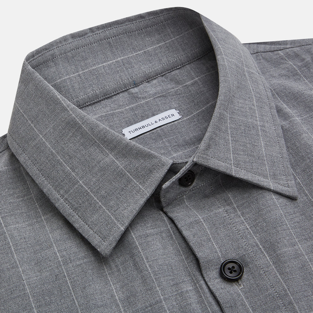 The Turnbull & Asser Grey Check Hampstead Overshirt features a light gray check pattern, pointed collar, chest pocket, and two zippered side pockets, displayed flat against a white background.