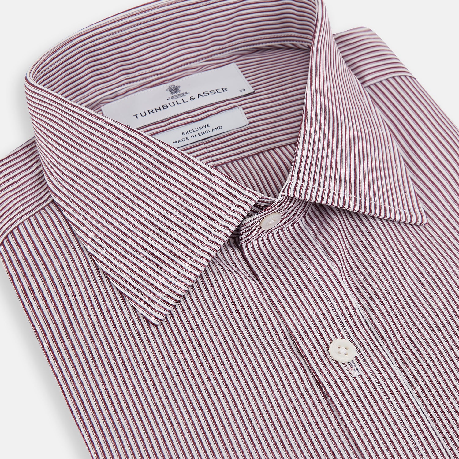 View 2 of product The Turnbull & Asser Burgundy Shadow Stripe Mayfair Shirt is a long-sleeved button-up with thin vertical burgundy stripes, classic collar, and buttoned cuffs, shown laid flat on a white background.