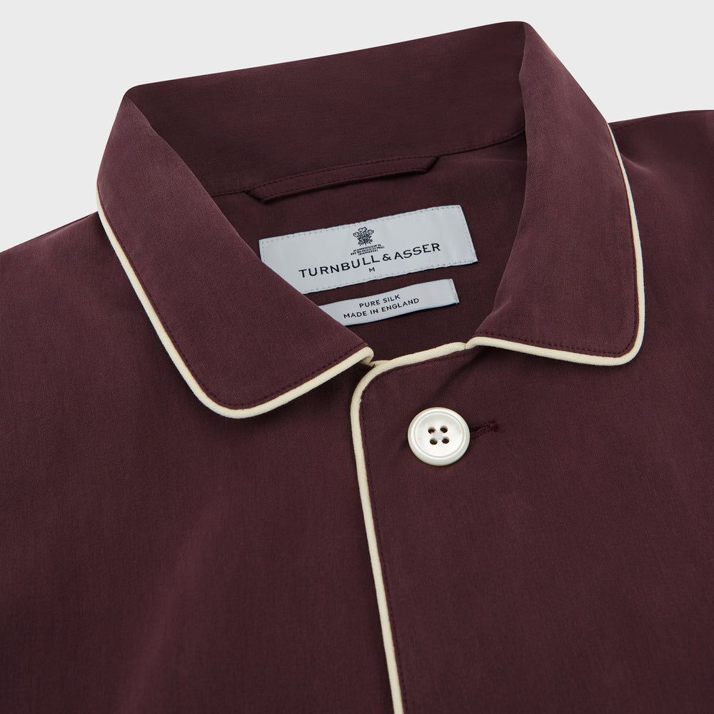The Turnbull & Asser Plain Plum Silk Modern Pyjama Set, made in England, features white piping, a button-up long-sleeve top with three pockets, and matching pants. The pyjamas are shown against a white background.