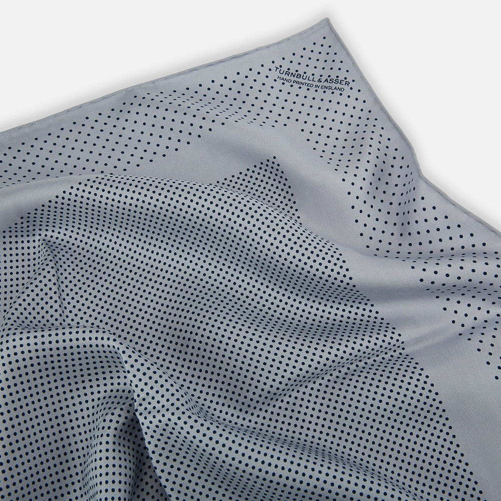 The Turnbull & Asser Grey Dot Silk Pocket Square features a light gray silk design with navy polka dots and a solid border. Elegantly handmade, it is shown with one corner folded to display both sides.