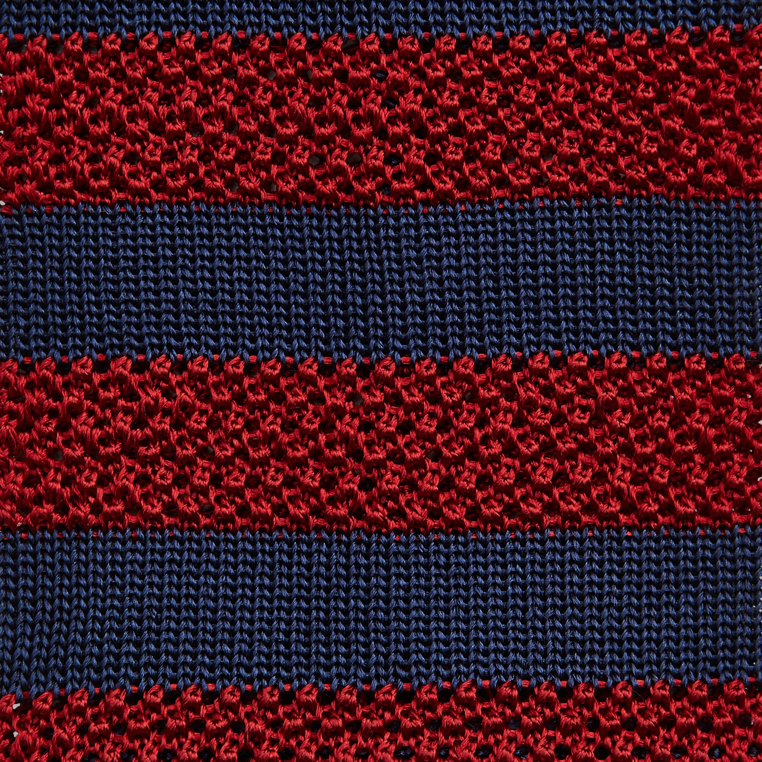 View 3 of product The Turnbull & Asser KNITTED STRIPE RED TIE features bold red and navy wide stripes in a rolled-up, knitted design—an eye-catching accessory shown on a plain white background.