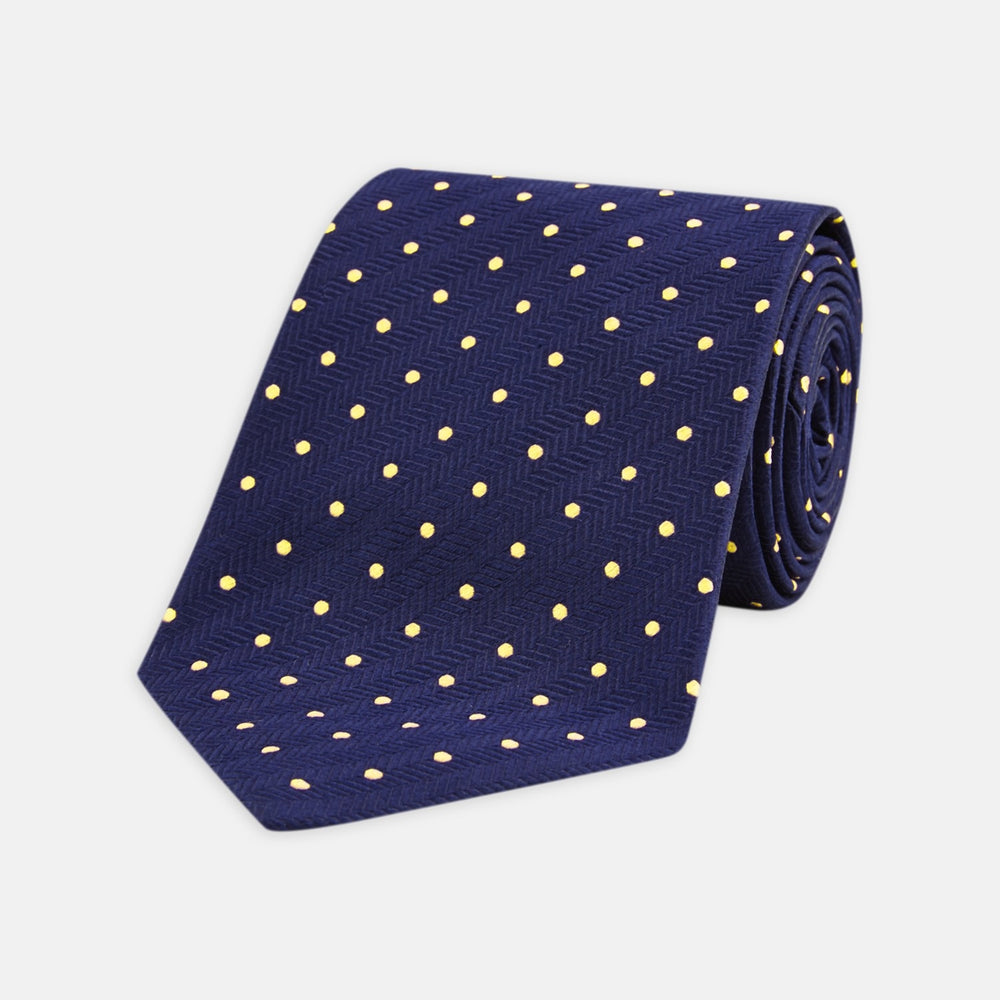 The Turnbull & Asser Navy Spot Woven Herringbone Silk Tie features a navy blue base with evenly spaced yellow polka dots, making it an essential accessory for smart attire. Displayed rolled on a white background.