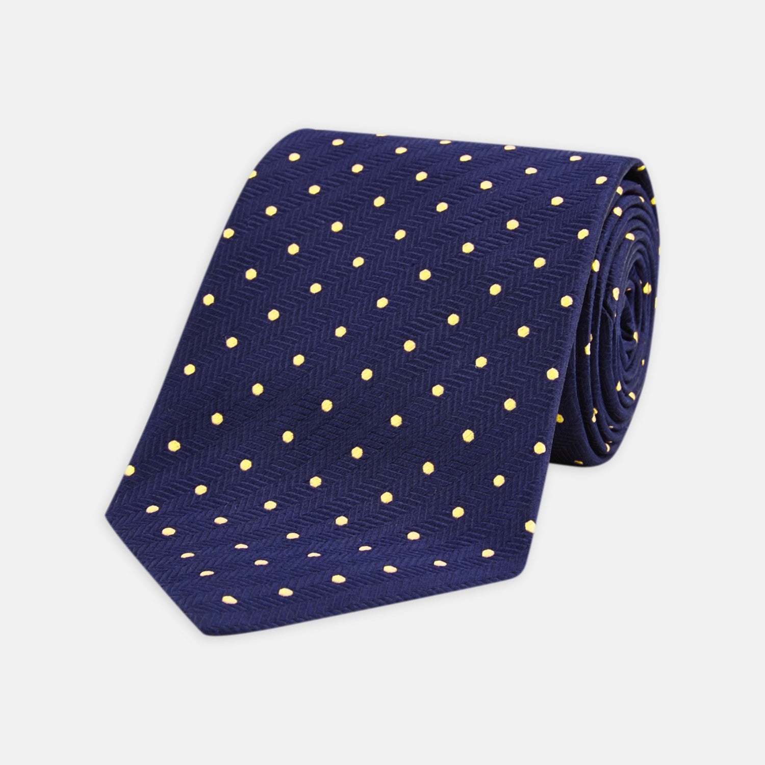 View 1 of product The Turnbull & Asser Navy Spot Woven Herringbone Silk Tie features a navy blue base with evenly spaced yellow polka dots, making it an essential accessory for smart attire. Displayed rolled on a white background.