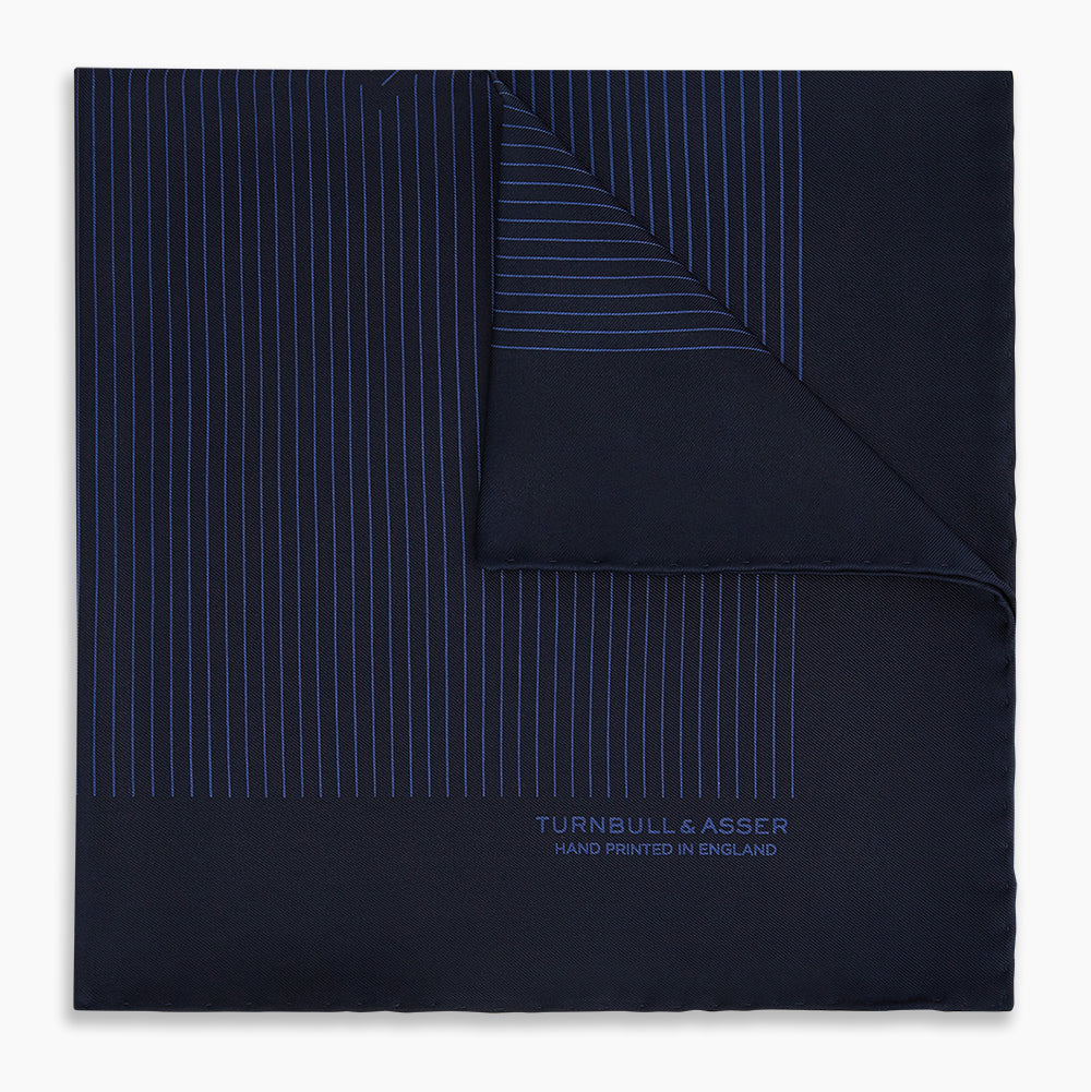The Navy Stripe Silk Pocket Square by Turnbull & Asser features a dark navy silk with thin light blue stripes and hand-rolled edges, folded to reveal both sides and “TURNBULL & ASSER HAND PRINTED IN ENGLAND” in the corner.