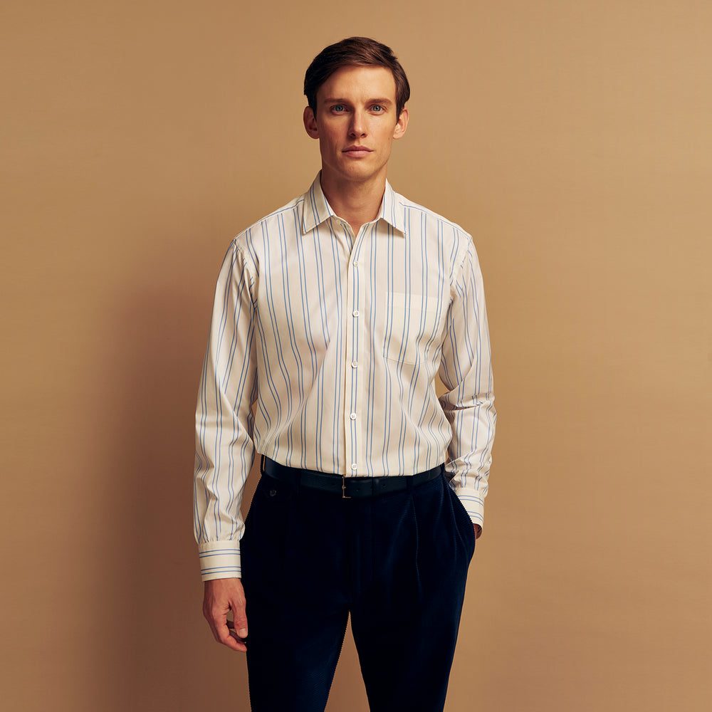 A man with short brown hair stands against a beige background in a Turnbull & Asser Blue and Off-White Stripe Cotton Weekend Fit Finch Shirt, featuring vertical stripes and a Derby collar, paired with dark pants. One hand is in his pocket; he has a neutral expression.