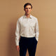 A man with short brown hair stands against a beige background in a Turnbull & Asser Blue and Off-White Stripe Cotton Weekend Fit Finch Shirt, featuring vertical stripes and a Derby collar, paired with dark pants. One hand is in his pocket; he has a neutral expression.