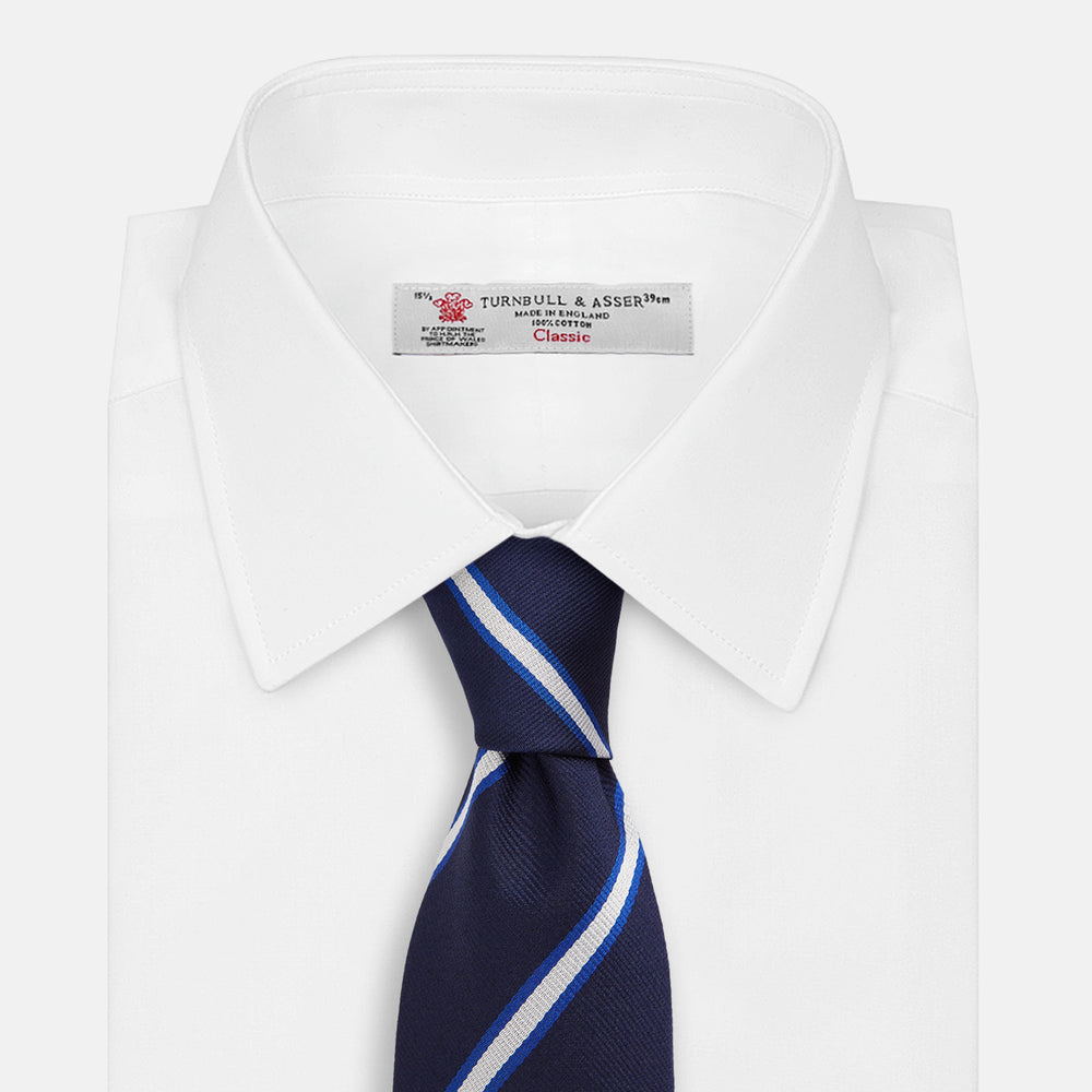 The White and Navy Blazer Silk Tie by Turnbull & Asser features diagonal white and light blue stripes on navy silk, shown rolled on a white background. A refined choice to complement any office shirt.