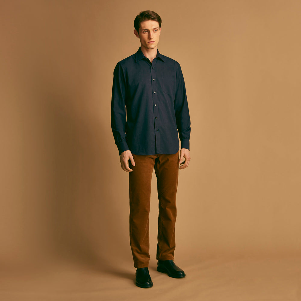 A man stands against a beige background wearing the Turnbull & Asser Navy Weekend Fit Cotton & Wool Shirt with a Derby collar and 1 button cuffs, paired with brown pants and black shoes, looking to the side with a neutral expression.