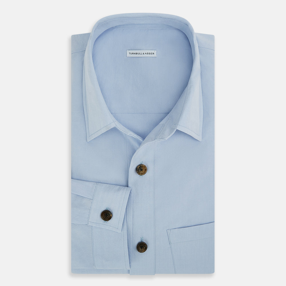 The Turnbull & Asser Pale Blue Hyde Overshirt is a long-sleeve, sanforised cotton button-up with a single chest pocket and dark buttons, shown on a plain white background.