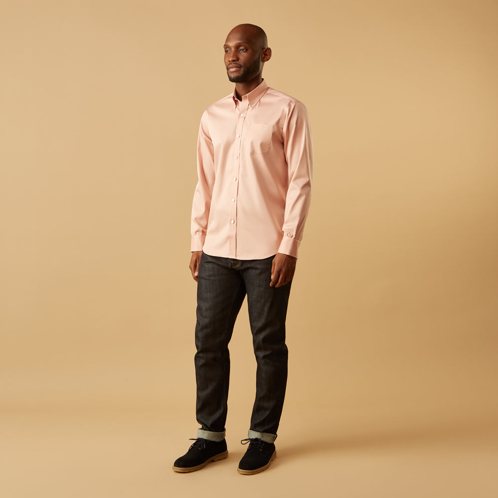 A man faces forward in a relaxed pose against a beige background, wearing the Turnbull & Asser Pink Piece Dyed Weekend Fit Suffolk Shirt with a button-down collar, dark cuffed jeans, and black shoes.