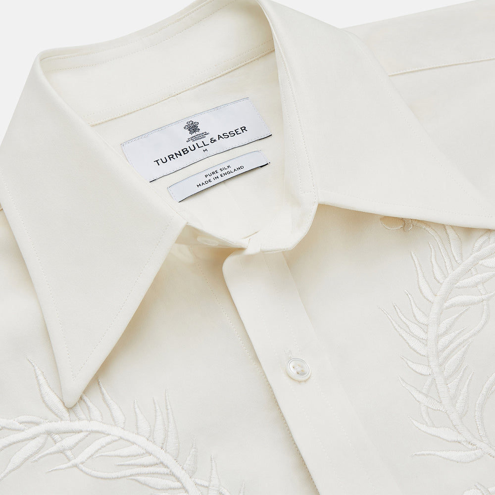 The Cream Silk Long Point Embroidered Eamon Shirt by Turnbull & Asser features long sleeves, a pointed collar, and delicate botanical embroidery. Its buttoned cuffs and tailored fit make it an elegant partywear choice.