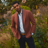 View 1 of product A man stands in a lush garden, hands in pockets and relaxed, wearing the Turnbull & Asser Red Lambswool Herringbone Bern Blazer, a striped dress shirt, patterned scarf, and dark trousers.