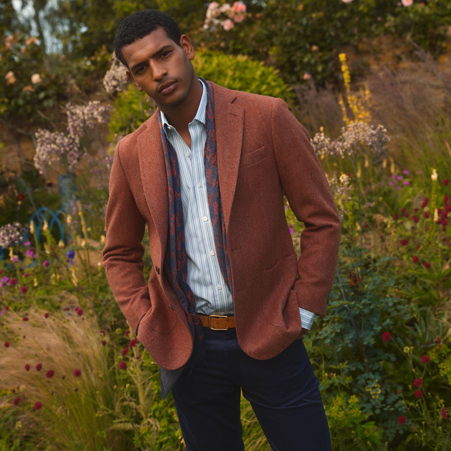 View 1 of product A man stands in a lush garden, hands in pockets and relaxed, wearing the Turnbull & Asser Red Lambswool Herringbone Bern Blazer, a striped dress shirt, patterned scarf, and dark trousers.