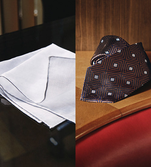 A white handkerchief with dark edges is neatly folded on a dark surface beside a patterned tie with geometric designs, displayed on a wooden table with a red chair nearby.