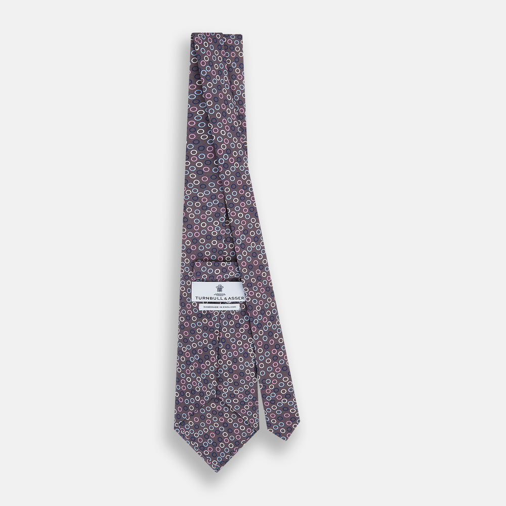 The Turnbull & Asser Purple and Blue Circle Ash Silk Tie features small blue, pink, and white circles on a dark purple background, adding an elegant touch when displayed on a light surface.