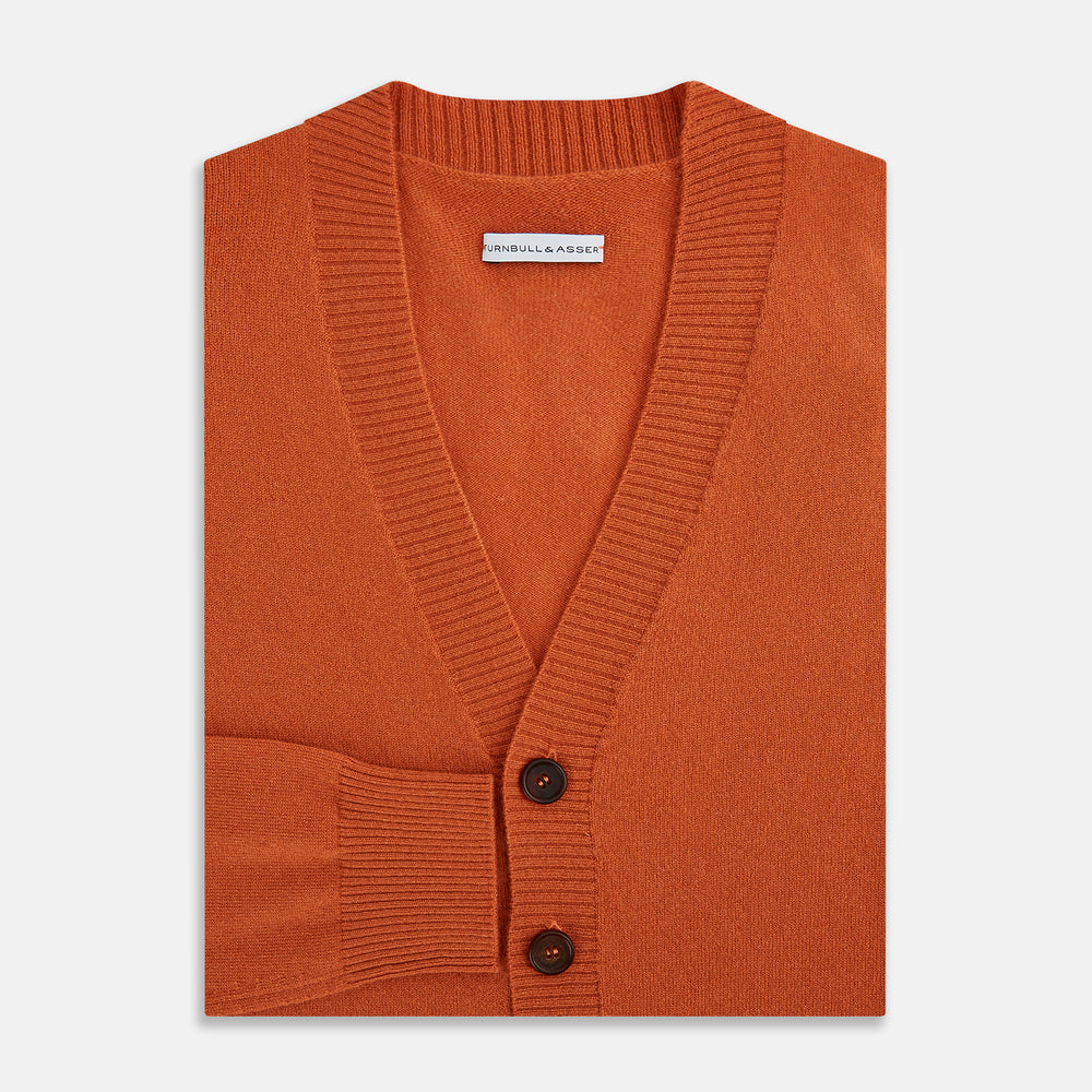 The Orange Cashmere Cardigan by Turnbull & Asser showcases vibrant burnt orange, a V-neck, dark buttons, ribbed cuffs and hem, long sleeves, and two front pockets. Part of the knitwear collection; displayed on a white background.