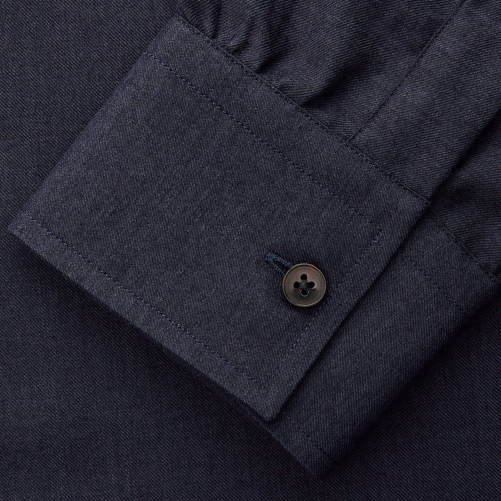 A man stands against a beige background wearing the Turnbull & Asser Navy Weekend Fit Cotton & Wool Shirt with a Derby collar and 1 button cuffs, paired with brown pants and black shoes, looking to the side with a neutral expression.