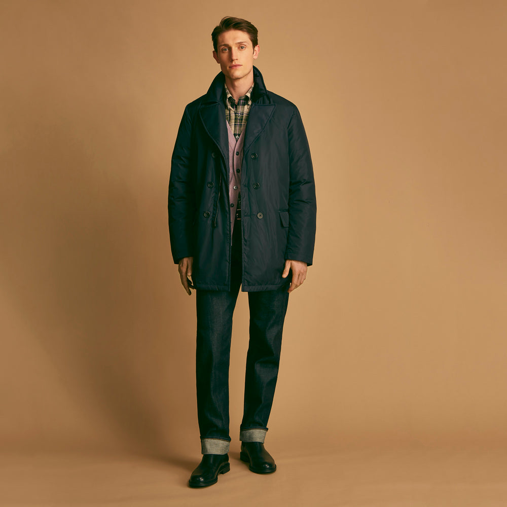 A man stands against a beige background, wearing the Turnbull & Asser Navy Burrows Double Breasted Coat over a plaid shirt and mauve cardigan, paired with dark rolled-up jeans and black shoes, looking directly at the camera with a neutral expression.