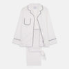 View 1 of product The Turnbull & Asser Women's Harriet Plain White Cotton Pyjama Set features long sleeves, black piping, a chest pocket, and matching pants with elastic waistband and ribbon tie, all displayed on a white background.
