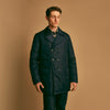 View 4 of product A man stands against a beige background, wearing the Turnbull & Asser Navy Burrows Double Breasted Coat over a plaid shirt and mauve cardigan, paired with dark rolled-up jeans and black shoes, looking directly at the camera with a neutral expression.