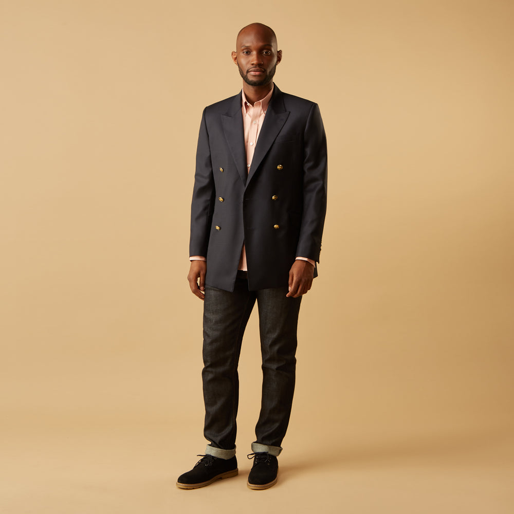 A man faces forward in a relaxed pose against a beige background, wearing the Turnbull & Asser Pink Piece Dyed Weekend Fit Suffolk Shirt with a button-down collar, dark cuffed jeans, and black shoes.