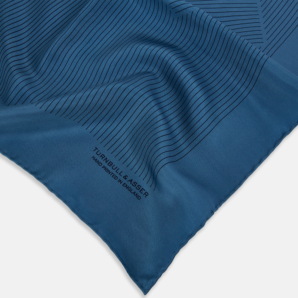 The Blue Stripe Silk Pocket Square by Turnbull & Asser features thin vertical lines, British heritage details, and "TURNBULL & ASSER HAND PRINTED IN ENGLAND" printed on the fabric, with one corner folded to reveal the reverse side.