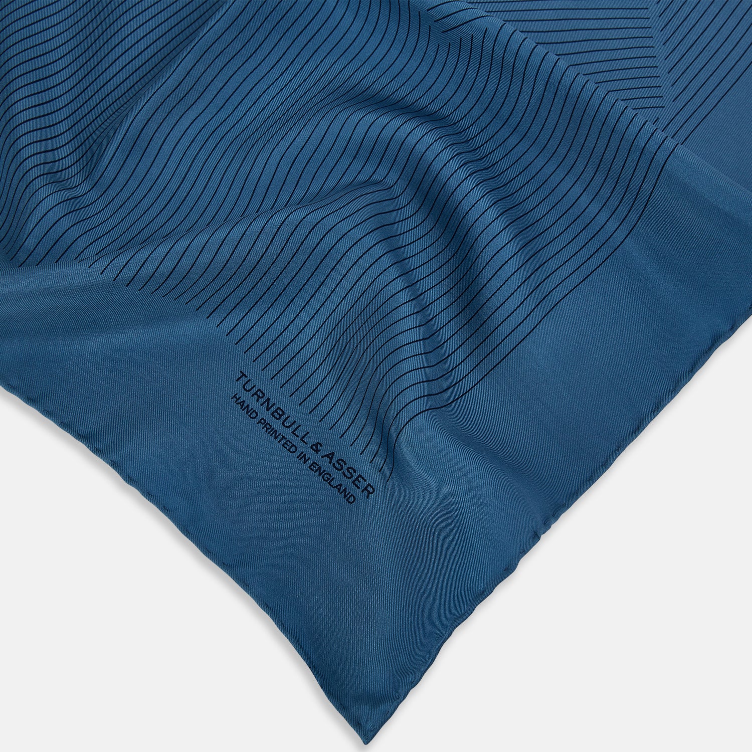 View 2 of product The Blue Stripe Silk Pocket Square by Turnbull & Asser features thin vertical lines, British heritage details, and "TURNBULL & ASSER HAND PRINTED IN ENGLAND" printed on the fabric, with one corner folded to reveal the reverse side.