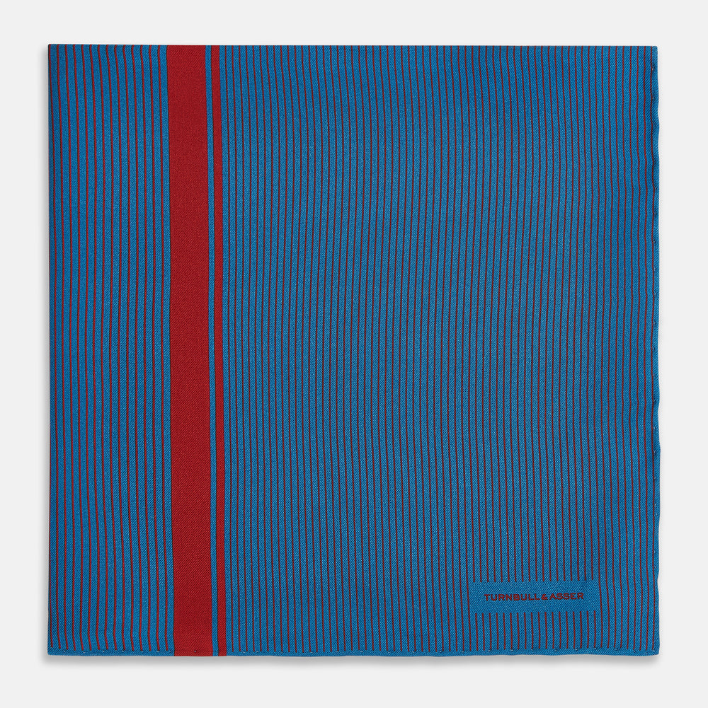 A blue silk pocket square by Turnbull & Asser, featuring thin black stripes, a striking vertical red stripe, and a turned-over corner that reveals the reverse side. The brand name is printed discreetly in one corner.