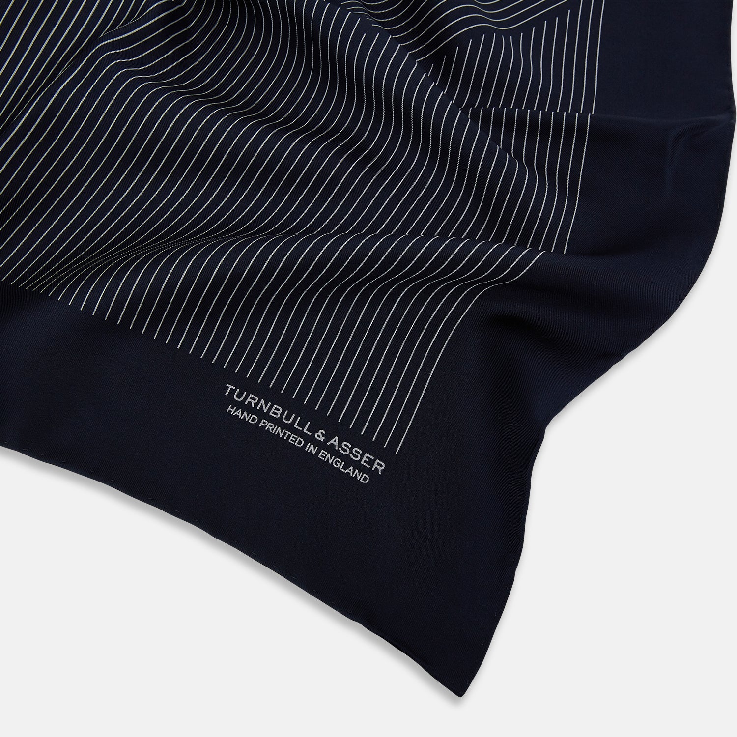 View 2 of product A Navy and Grey Stripe Silk Pocket Square by Turnbull & Asser, featuring hand-rolled edges and brand text in the corner. One corner is folded to show the reverse side.