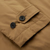 View 7 of product A person wears the Turnbull & Asser Camel Quintern Sealup Raincoat with Detachable Liner, paired with dark pants, standing against a plain light gray background, showing the sleek technical rainwear style with one hand in a pocket.