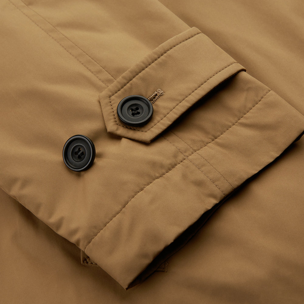 A person wears the Turnbull & Asser Camel Quintern Sealup Raincoat with Detachable Liner, paired with dark pants, standing against a plain light gray background, showing the sleek technical rainwear style with one hand in a pocket.