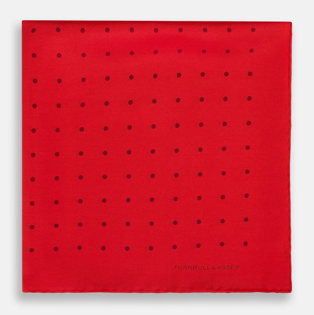 The Turnbull & Asser Red and Burgundy Spot Silk Pocket Square features evenly spaced small spots and the brand logo in the bottom right corner—perfect for formal wear. One corner is folded to show the reverse side.